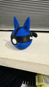 En Poké Ball-inspireret kugle i blå og sort med spidse ører, der giver et legende og karakteragtigt udtryk. Den har en glat finish og en frontknap, som tilføjer et interaktivt og stilrent design.