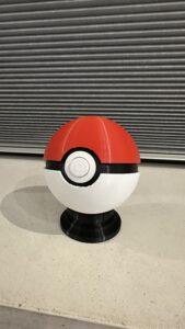En klassisk Poké Ball i rød, hvid og sort, placeret på en stabil base for præsentation. Designet er rent og ikonisk med veldefinerede proportioner og en flot, blank overflade.